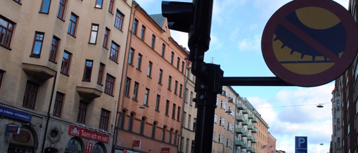 Förbudsmärke i stadsmiljö som indikerar förbud mot dubbdäck. Bakom skylten syns flerbostadshus och butiker.