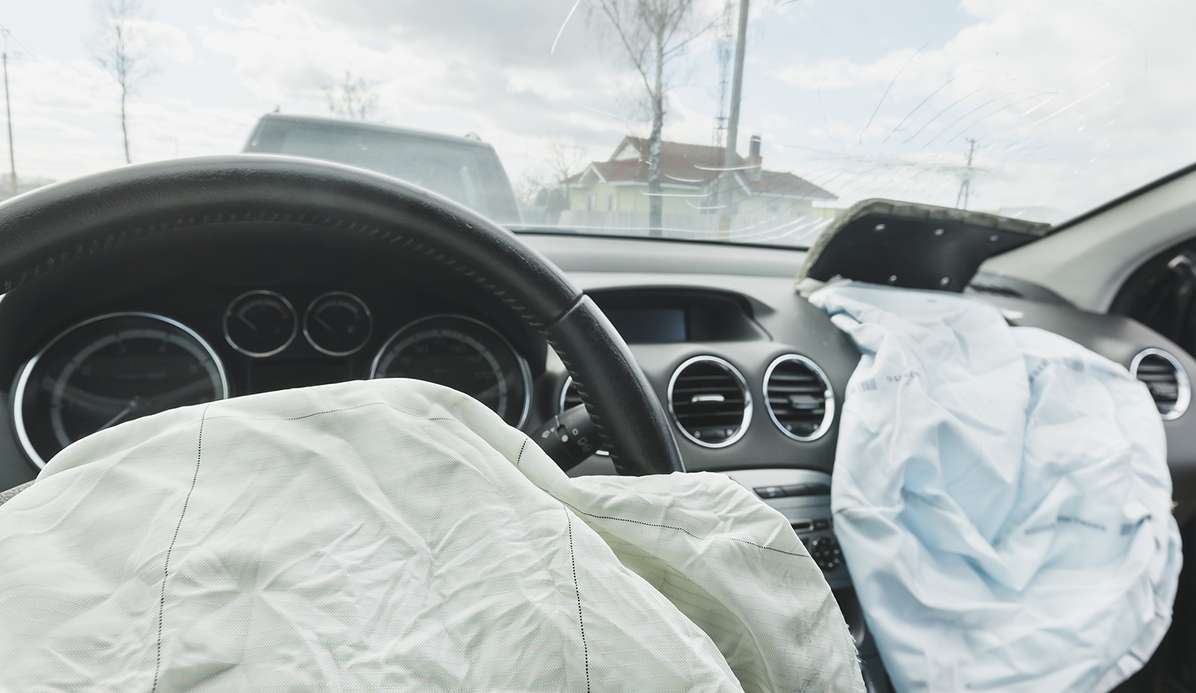 Bilinteriör med utlösta krockkuddar på ratten och passagerarsidan, med en sprucken vindruta och ett annat fordon synligt utanför.