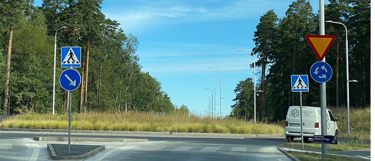 Rondell i ett grönskande område med vägskyltar för övergångsställe, påbjuden körriktning och väjningsplikt