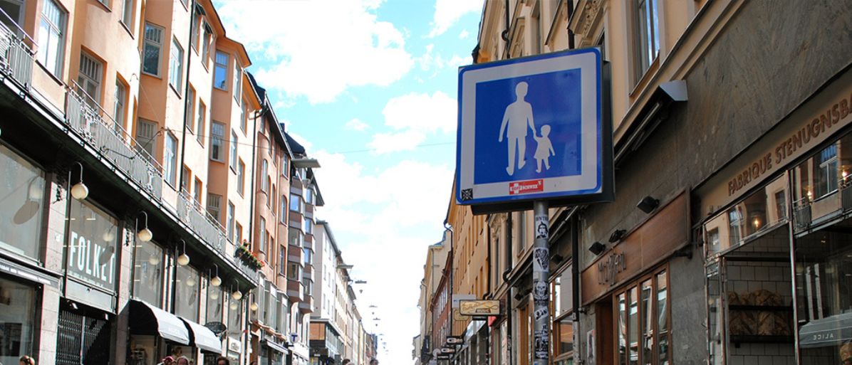 Skylt för gångfartsområde i en livlig stadsgata med butiker och bostäder.